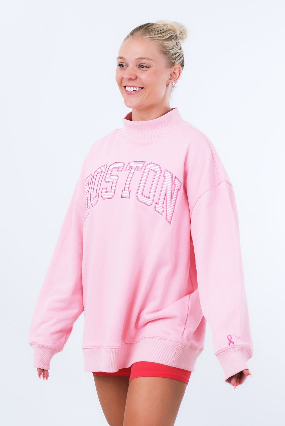 A woman modeling an oversized light pink sweatshirt with 'BOSTON' printed in arched pink letters across the chest, paired with red athletic shorts, white socks, and white sneakers against a white studio background.