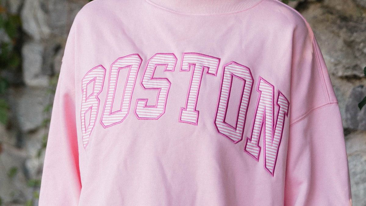 A person wearing a light pink sweatshirt with 'BOSTON' arched across the chest in outlined block letters, paired with light blue jeans, standing outdoors against a stone wall.