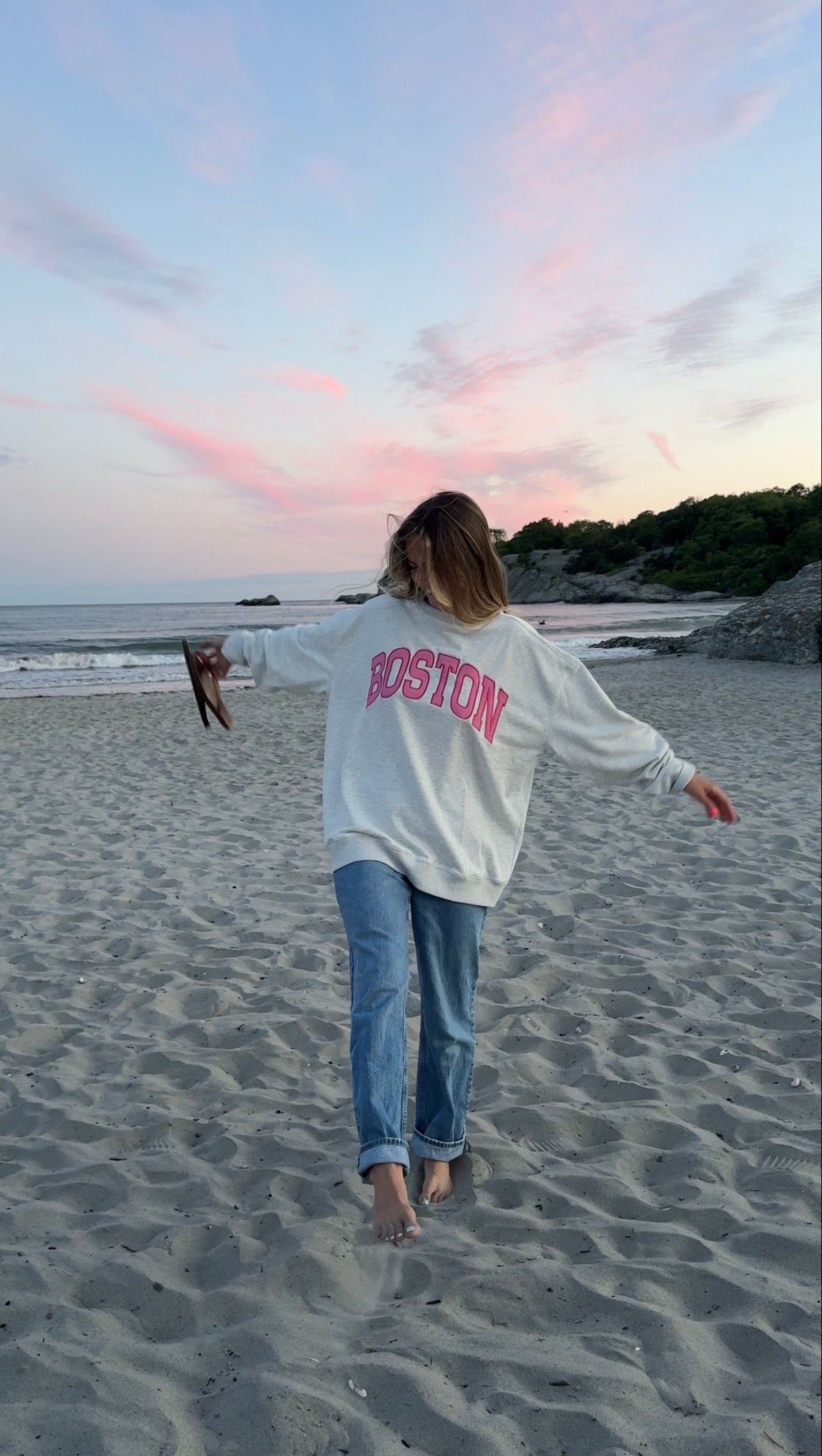 A woman stands on a sandy beach at sunset wearing a white sweatshirt with 'BOSTON' printed in pink across the back, paired with blue jeans.