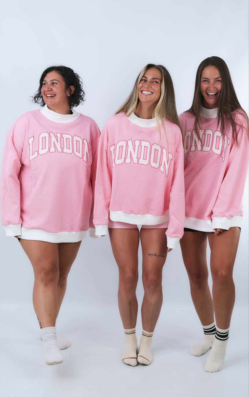 Three women wearing pink sweatshirts with 'London' printed on them against a white background.