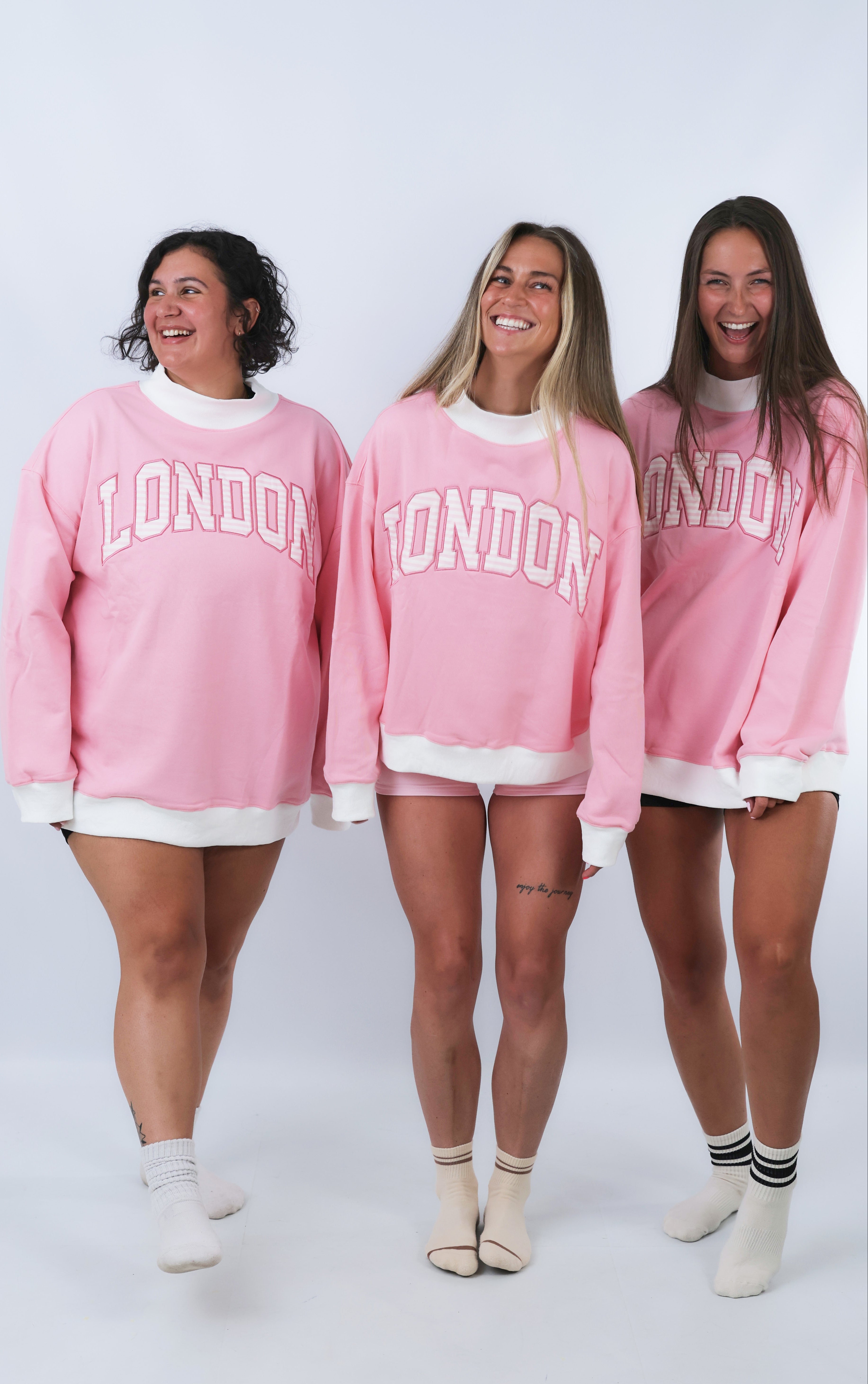 Three women wearing pink sweatshirts with 'London' printed on them against a white background.