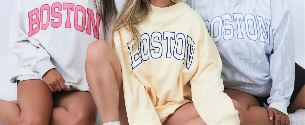 Three women sitting on the floor wearing oversized sweatshirts with 'BOSTON' printed across the chest in different colors: pink, navy, and blue. The photo is taken against a seamless white studio background.